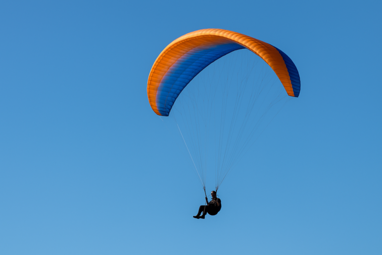 Paraglider voando em céu azul limpo com vela laranja e azul.
